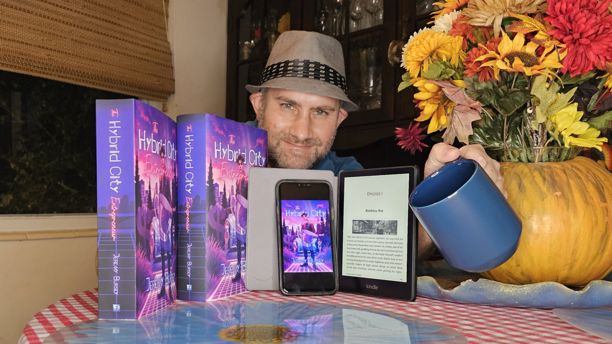 A man in a hat sits at a table with three books, an e-reader, a smartphone displaying a Jeremy Bursey book cover, a blue mug, and a vase of artificial flowers. A man in a hat sits at a table with three books, an e-reader, a smartphone displaying a Jeremy Bursey book cover, a blue mug, and a vase of artificial flowers.